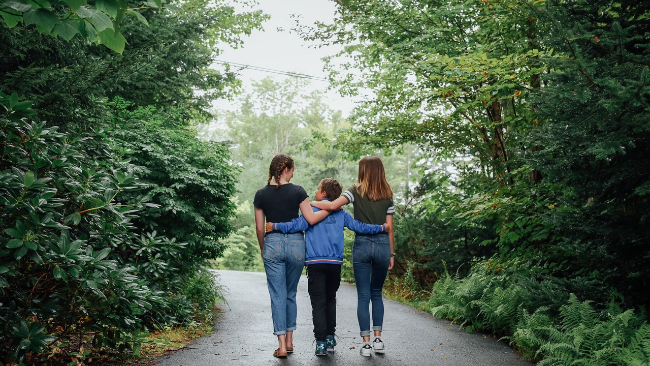 Three children linked arms walking away up a tree-lined driveway